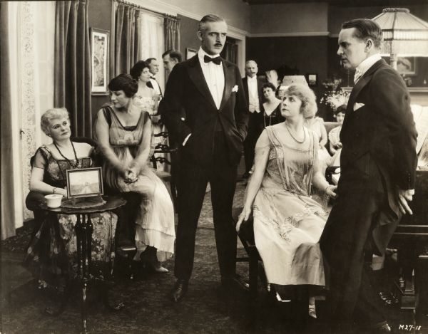 At a social gathering, Jack Fyfe (played by Wallace Reid in a tuxedo) glares at Walter Monahan (Joe King in white tie and tails) as Stella Barton (Kathlyn Williams at the piano) worriedly watches. Nine extras in evening dress are also in this scene still for the silent drama "Big Timber."