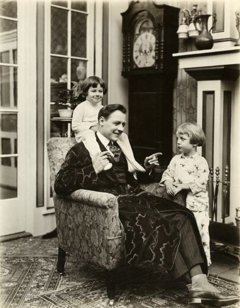 Harold Lockwood, as John Tremaine, Jr., sits in an overstuffed chair with one young actor on his shoulders and actor Ben Alexander at his feet, in a scene still for the Yorke Film Corporation silent drama "The Big Tremaine."
