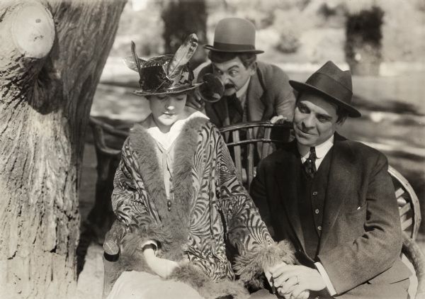 The stenographer Ethel (played by Fay Tincher) sits on a bench holding hands with Ed (Edward Dillon) as a man in a moustache, probably Ed's father (Max Davidson), examines her closely with a magnifying glass. This is a scene still for "Ethel Gets Consent," which was number 17 in the one-reel silent comedy series "Bill, the Office Boy."