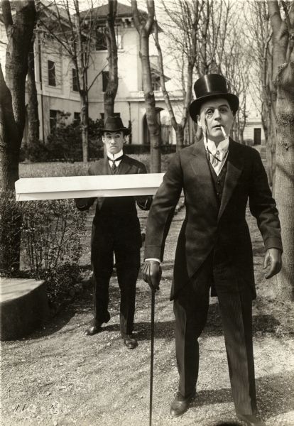 J. Courtleigh Brice (a spoiled son of wealth played by Harry Benham wearing a monocle) staggers down a path followed by a servant carrying a long box (Claude Cooper) in a scene still from the Thanhouser silent comedy "Daughter of Kings."
