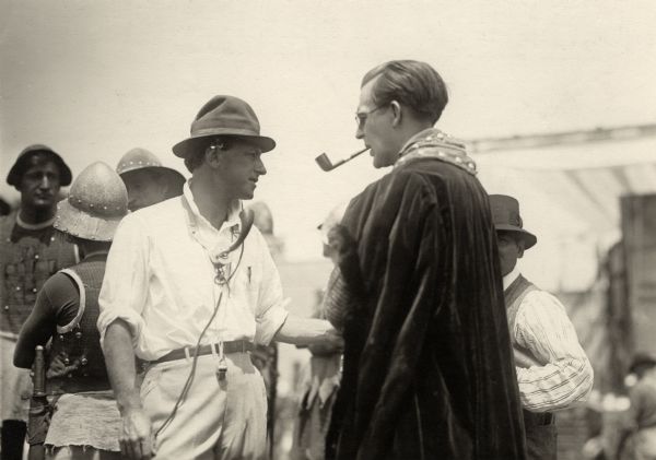 Director Cecil B. DeMille confers with actor Wallace Reid on an outdoor set for the 1916 silent drama "Joan the Woman." DeMille wears a chest microphone like those used by telephone operators. Reid is costumed in a velvet cloak and is smoking a pipe. Behind DeMille are three extras dressed as 15th century soldiers.