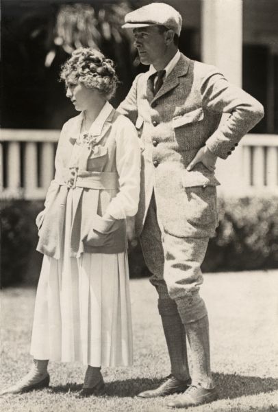 Publicity Still of Mary Pickford and Owen Moore Outdoors Silent film star couple Mary Pickford and Owen Moore pose on a lawn outdoors. She wears a loose, wide belted jacket over a white pleated dress. He wears a herringbone worsted wool suit with large patch pockets, plus fours, and a cloth cap.