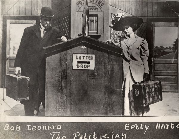 Robert Z. Leonard and Betty Harte stand on opposite sides of a post office writing desk in a scene still from a Selig Polyscope silent film, c. 1908.
