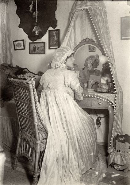 Actress Florence Lawrence seated at the vanity mirror in her dressing room wearing a white satin dressing gown and night cap.