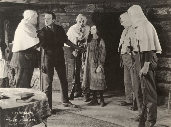 In a log cabin, Bradley Yates (played by Frank Mayo) is confronted by four armed vigilantes, who wear klan-style hoods. The mountain girl Talithy Millicuddy (played by Mary Philbin in a gingham dress) watches with interest.