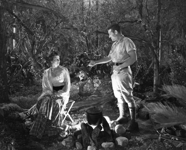 Tom Dekker (played by Tom Neal) and Teresa (Carole Matthews) talk by the fire at their camp in the Amazon jungle.