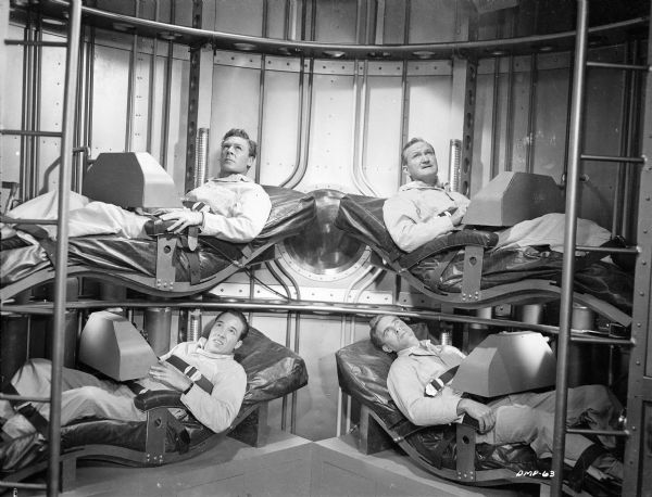 Astronauts in their bunks in a scene still for "Destination Moon." Four astronauts are strapped into their bunks in a spaceship interior. From top left, proceeding clockwise, the actors are John Archer, Tom Powers, Warner Anderson, and Dick Wesson. All are looking up with worried expressions.