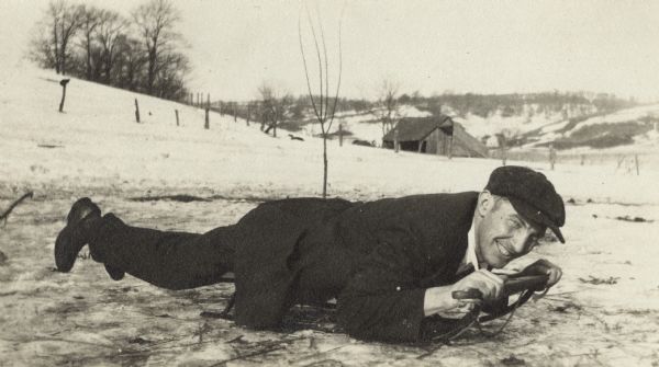 Winter scene with a man lying on a small sled with a big smile on his face. He is wearing dark clothes and a cap. A snow-covered hill, a barn and fences are in the background.