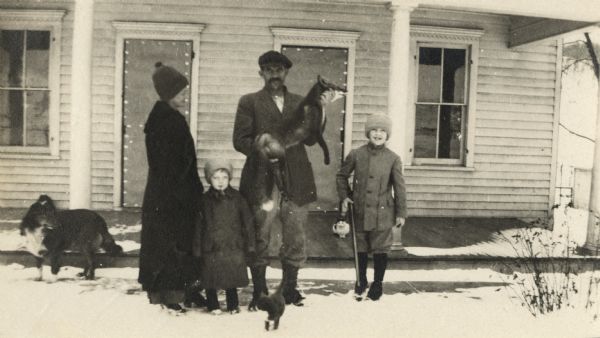A family poses outdoors in the snow in front of their house. The father proudly holds a dead fox. The mother and youngest child stand on the left, and the older child stands to the right holding a gun. There is a cat at their feet and another on the porch. A dog is on the left.