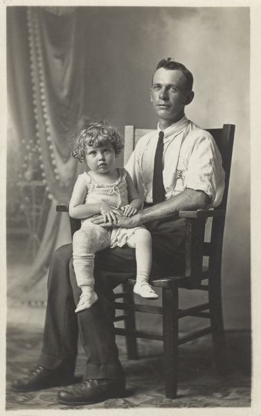 Full-length studio portrait in front of a painted backdrop of Charles Faust, seated in a chair, holding Lynne Faust Hilgers on his lap. He is wearing a shirt, tie, slacks, suspenders and shoes. She is wearing a little dress, stockings and shoes. She appears to have her right leg in a cast that reaches up to her hip.