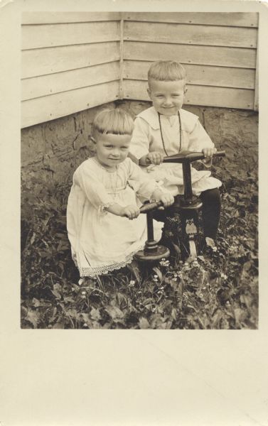 Two children pose outdoors in the corner of a yard next to a building. They are sitting on tricycles or pushbikes, one has an owl logo on the front. Both are wearing light-colored clothing and have identical haircuts. Foliage and flowers are at their feet.