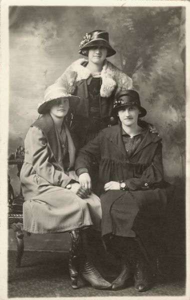 Full-length studio portrait of three women in front of a painted backdrop. Two women are seated, the third woman is standing behind and has her hands on their shoulders. All three women are wearing jackets over dresses, and hats decorated with flowers and ribbons. The standing women wears a fur cape. Two women are wearing watches or bracelets.