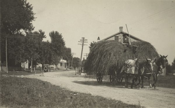 Photographic postcard of a haywagon pulled through a village by two horses. The driver is perched on top of the load. One horse wears a blanket with printing on it. Buildings, trees, an automobile and power poles are in the background. The large building with a front porch and a balcony has a sign that reads: "Undertaker."