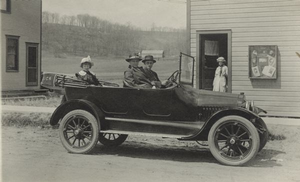 Photographic postcard of a man, woman and young girl posing in their Buick convertible in front of the Matthew Witt Photographic Studio. Another woman stands in the open doorway of the studio. Everyone is wearing a hat. Samples of the studio's work are on display in a case next to the door. Another building is on the left, and a farm building and tree-covered ridge are in the far background.