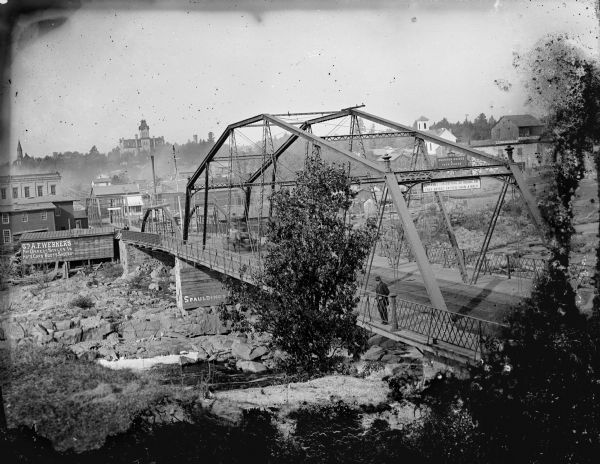 Harrison Street Bridge View looking down hill of bridge, probably the Harrison Street Bridge, leading into town over the river. There is a man standing on the bridge, and a blurred images of two men in a horse-drawn vehicle. An industrial building on the far rocky shoreline has a sign that says: "Go To A.F. Werner's for correct styles in Hats Caps Boots Shoes, Etc."