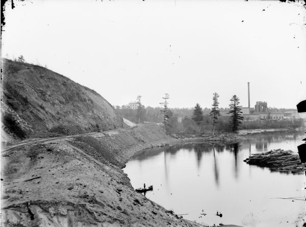 View down hill of man posed sitting in a boat near the bank of a river taken from an iron mound of the York Iron Works. There is a view of the blast furnace and the boarding house.