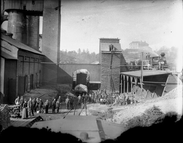 Elevated view of York Iron works. Large group of men posing in yard. The boardinghouse is on a hill in the far background. Early iron was mined at Mayville and Iron Ridge in Dodge County. Early foundries did business in castings, agricultural machinery and blacksmithing.