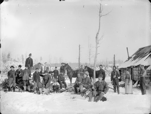 Group of men, a dog and one woman posed standing and sitting in the snow-covered yard near a wooden building,. Three men hold rifles, two men display horses, while another displays a team of two horses.	