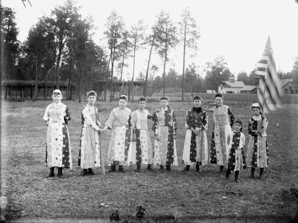 Nine girls are posing standing and wearing patriotic costumes. One girl is holding a flag of the United States. Probably a smaller group, from a total of twenty-eight girls, dressed in red, white and blue costumes, drilled by J.R. Ogden for a Memorial Day honor guard in 1904. Based on information in the "Melrose Chronicle," May 23, 1984, citing an article of April 28, 1904.	