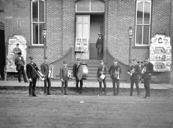 Brass band with eight musicians posed standing on the street in front of the Opera House. At the far right is Carl Pope. Signs advertise "John T. Hinds in Ireland As Tt Was." Two men stand in the open doorway of the Opera House entrance, and two boys stand on the sidewalk.
