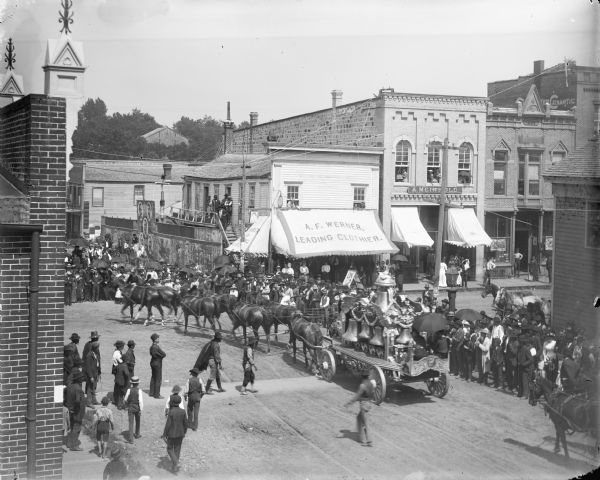 Elevated view of a crowd gathered at the intersection of First and Main Streets to watch a bell wagon pass in a circus parade, probably the Ringling Brothers Circus. The bell wagon has been owned by the Circus World Museum since 1985.	