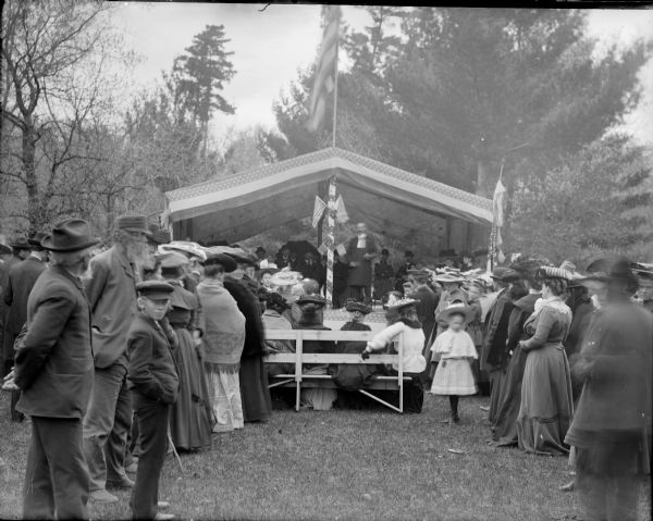 Crowd of men, women, girls and boys gathered around a tent, probably listening to a political or patriotic speech or debate.	