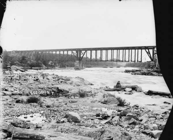 View from shoreline of two women posed on logs on rocky shoreline by the river. Four or five young boys sit on the rocks on the left. There is a railroad bridge is in the background. Graffiti letters painted on a rock nearby say, "Sozodon T." There are wooden structures in the river beyond the bridge, perhaps sorting piers for log transportation.	