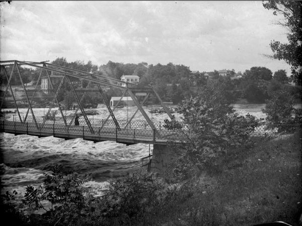 Elevated view from shoreline of high water passing under a bridge. There is a woman standing at the railing on the bridge. The town is on the far shoreline in the background. A sign on the bridge says: Clinton Bridge, Iron Works".