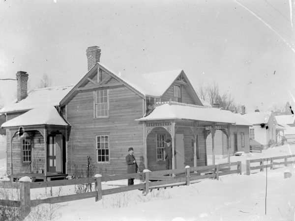 View from road of two men standing behind a fence next to a snow-covered frame house. A woman and young girl pose standing on the porch of a house two houses down on the right.