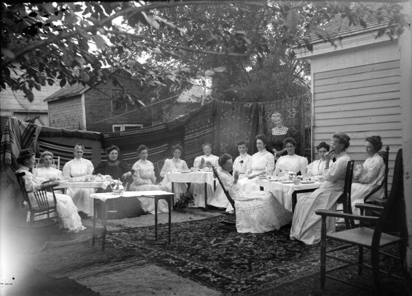 Group of fifteen women gathered around three tables behind a home, probably the DMC Club, an exclusive social and sewing society named for an imported brand of French fancywork cotton crocheting, also known locally as the Damn Mean Crowd Club by outsiders. Identified by Joanne Dougherty, sitting from left to right, as: unidentified, Mrs. Ludovic Dimmick, Mrs. Louis Jones, unidentified, Mrs. Anton Johnson, Mrs. Rufus Jones, Mrs. J.J. McGilivray, Flora LeClair (in rocking chair), unidentified, Mrs. John Marsh, unidentified, Edna Richards Turner, Mrs. Will Richards, Nina Mason Werner, and Mrs. Charles Van Schaick, standing. Behind them quilts are hanging on a line from the edge of the house all the way across the background, and there are rugs on the ground.