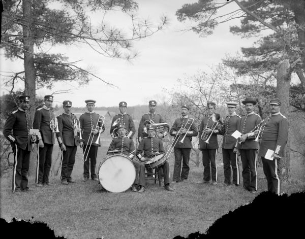 Eleven uniformed musicians holding their instruments stand in a semi-circle, with the two drummers seated with their drums. The band consists of 2 trombones, 3 trumpets, 2 coronets, 2 clarinets, 2 euphoniums, a snare drum and base drum with cymbal.