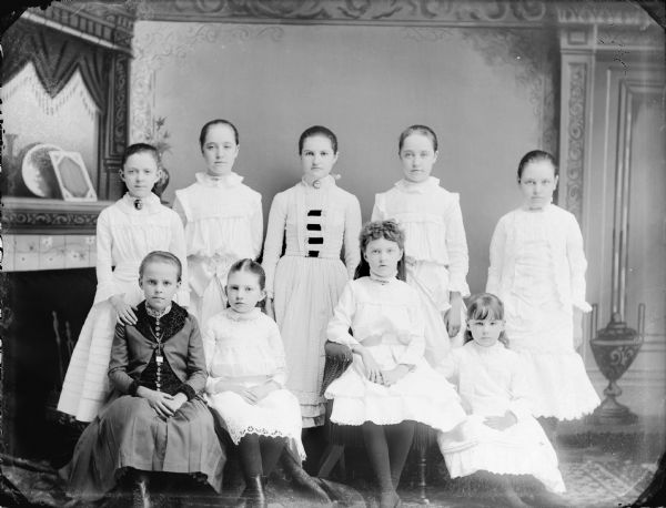 Studio Portrait of Nine Girls Studio portrait in front of a painted backdrop of nine girls, four posed sitting and five standing. Girls standing, from left to right, Julia Murray, Spaulding twin, Alice Mills, Spaulding twin, and unidentified. Second sitting girl from the left is Bessie Smith.
