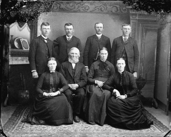 Studio portrait in front of a painted backdrop of an elderly man and woman posed sitting surrounded by four standing men and two sitting women. Probably the Nels Nelson family from Curran, Wisconsin.