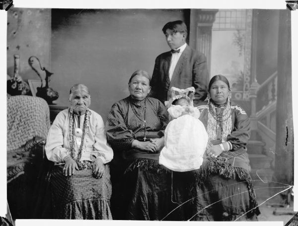 Five generation studio portrait of a Ho-Chunk family. Sitting (l to r) Great-Great-Grandmother, Nancy Brown; Great-Grandmother, Lucy Goodvillage, Blowsnake; Grandmother, Annie Blowsnake, Thundercloud. Standing: Adam Thundercloud; Baby: son of Adam Thundercloud. In the background is a painted backdrop.