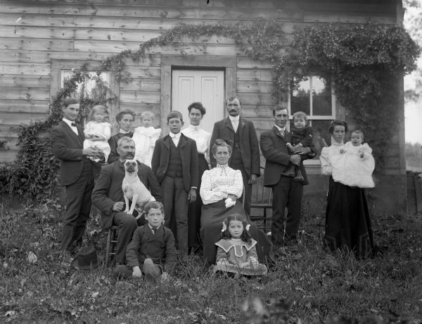 Group posing standing and sitting in the grass in front of a board house, possibly the Bill Jay family. The two children sitting are probably Edward and Sadie Ross.