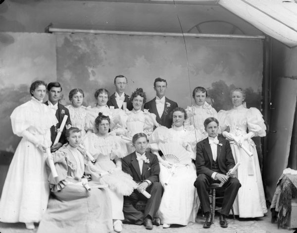 Studio portrait of two men and three women posing sitting, in front of three men and six women posing standing in front of a painted backdrop in a photographer's studio. The men and women are holding diplomas, probably the Black River High School Class of 1895.