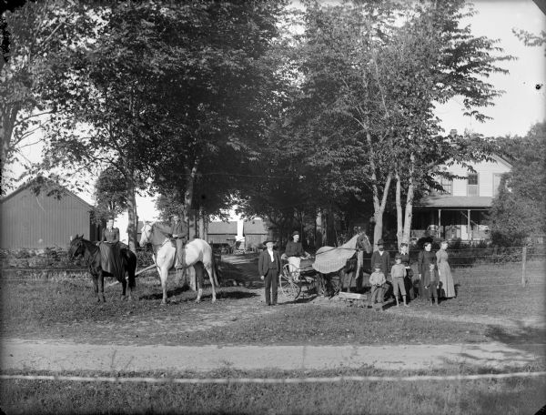View across road towards a group of people posing in front of a farm. On the left a man and woman are each sitting on separate horses, and in the center another man is sitting in a buggy pulled by a single horse wearing a blanket. Two more men, three women, and three boys are standing near the horses. The group is in the driveway, and behind them is a yard enclosed by a wire fence in front of a frame house and farm buildings partially obscured by trees.