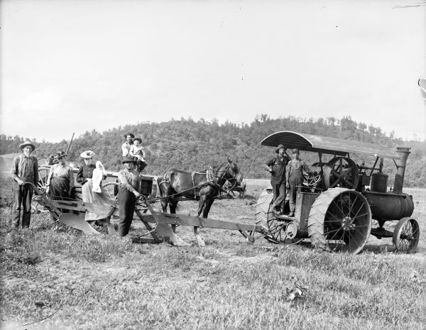 Two men are posing standing on a steam tractor on the right, probably a Linnell, attached to sod-breaking plows steered by two men on the left. Behind them are standing two women, one holding an infant. In the center background are a man and boy posing sitting in a wagon pulled by a team of two horses. In the far background are tree-covered hills.	