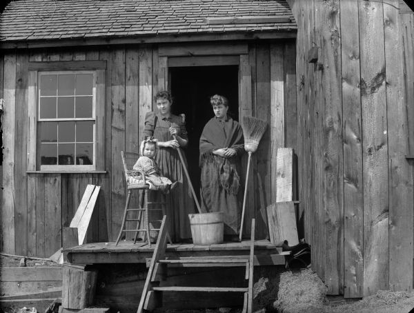 Two women are standing on the small porch outside an open kitchen door, near implements used for daily household tasks such as washing, scrubbing, sweeping, and child care. A young child is sitting in a high chair on the porch near a wooden bucket.