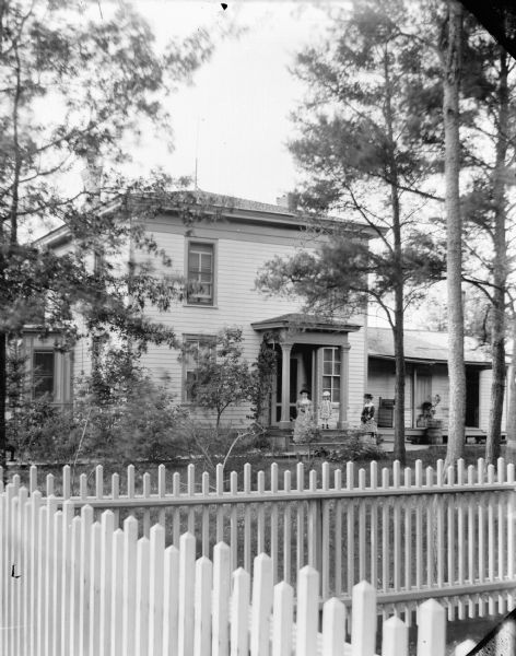 Two Women and Young Girl on Porch of Frame House View over fences towards two women and a young girl posing standing on the porch of a wooden frame house. The two women are wearing long dresses with lacy collars, and hats and gloves. The young girl is wearing a short, ruffled dress and a hat.