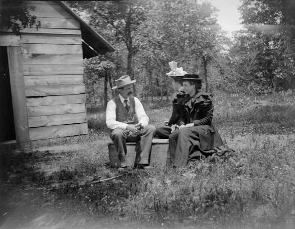 Man and Two Women Sitting Outdoors Group portrait of a man and two women posing sitting on a wooden box in a yard near the front of a board building.