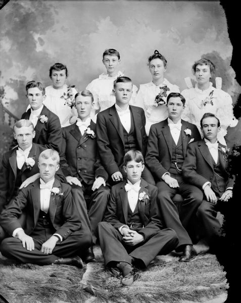 Group Portrait of Young Men and Women in Formal Attire Studio portrait in front of a painted backdrop of three men posing sitting on the floor, in front of five men posing sitting in chairs, and four women standing. Most of the men are wearing boutonnières, and the woman are wearing corsages. Possibly a graduation class.