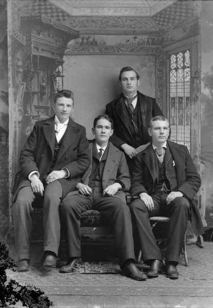 Anton Johnson, Fred Werner, Ben Bright and unknown man Studio portrait in front of a painted backdrop of four young European American men. They all wear coats over dark-colored suits. Three of the men sit in front, and one man stands behind them. Men identified from left to right as probably Anton Johnson, unidentified man, Fred Werner (standing), and Ben Bright.