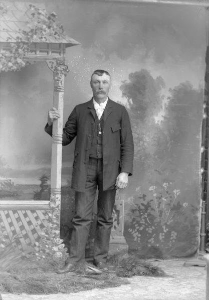 Portrait of Man in Gazebo Scene Full-length studio portrait in front of a painted backdrop of a European American man with a moustache posed standing with his right hand holding a prop post. He is wearing a dark-colored suit coat, vest, striped trousers, long light-colored bow tie, rings on both pinkies, and a watch chain. The edge of the backdrop is on the far right.