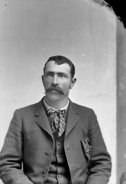 Portrait of a Man with Handlebar Moustache Studio portrait of a European American man with a moustache posed sitting. He is wearing a dark-colored suit coat, vest, and long striped bow tie.