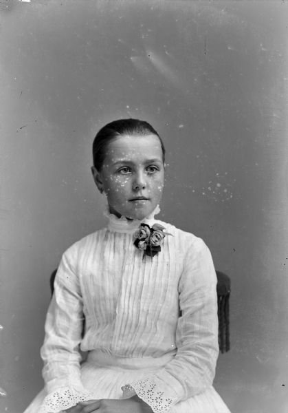 Studio Portrait of European American Girl Waist-up studio portrait of a young European American girl posed sitting in a chair with a tasseled back. Her hair is pulled back, and she is wearing a light-colored pleated dress with a high ruffled collar and lace trim at the wrists. She is also wearing earrings and a corsage.