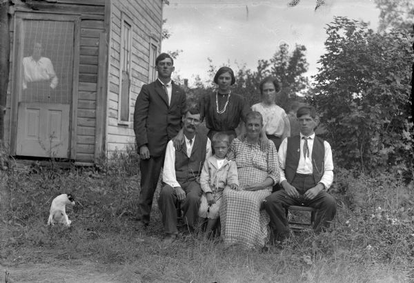 Outdoor Group Portrait of European American Family Outdoor group portrait of an unidentified family. An older man and woman are posing sitting in the front, with a child between them, and a younger man sitting on the right. Behind them are standing two women and a man. The women standing in the center has her arms around the older man and woman. Another person is standing behind the screen door of a house on the left. A dog is sitting in the grass to the left of the group.