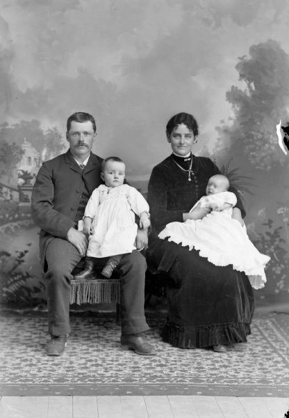 Studio Portrait of European American Family Studio portrait in front of a painted backdrop of an unidentified family. A man and a woman are posing sitting, both holding infants in their laps. Both infants are wearing white dresses.