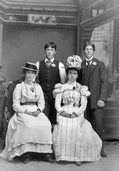 Studio Portrait of Two European American Couples Full-length studio portrait in front of a painted backdrop of two unidentified women wearing hats posing sitting, and two unidentified men posing standing behind them.