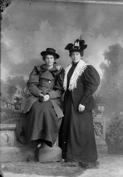 Studio Group Portrait of Two European American Women Studio portrait in front of a painted backdrop of an unidentified woman posing sitting on the left and an unidentified woman posing standing on the right.